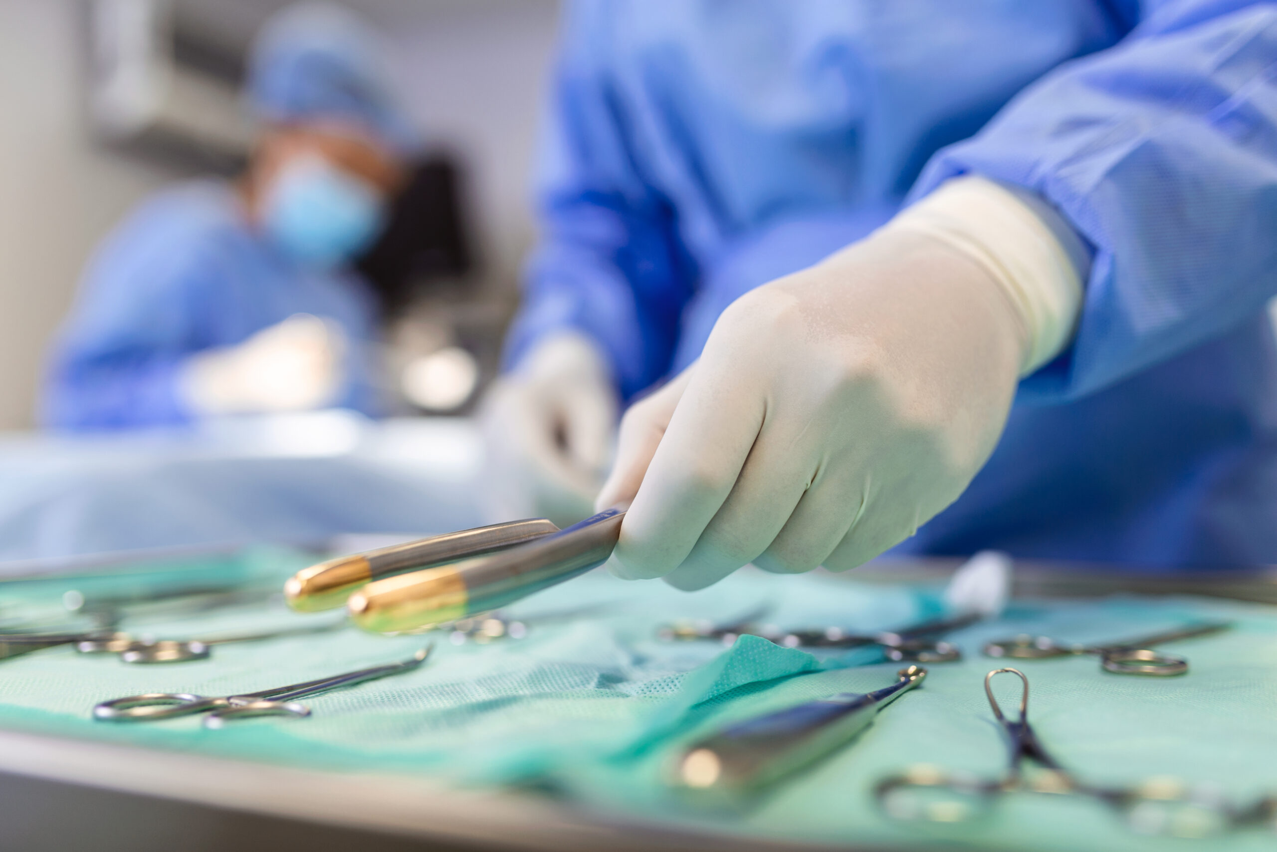 Nurse hand taking surgical instrument for group of surgeons at background operating patient in surgical theatre. Steel medical instruments ready to be used. Surgery and emergency concept